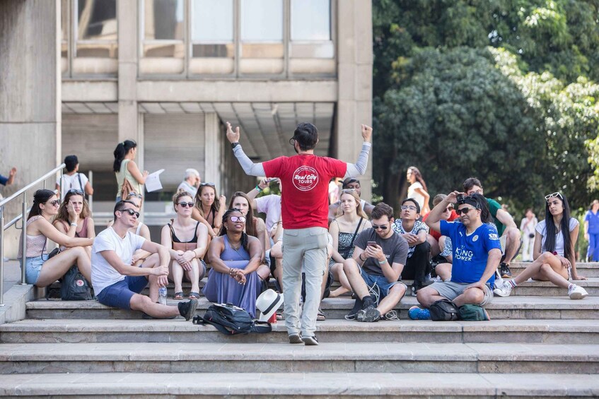 A person in a red shirt with a logo, standing on steps with arms raised, surrounded by seated people. 3