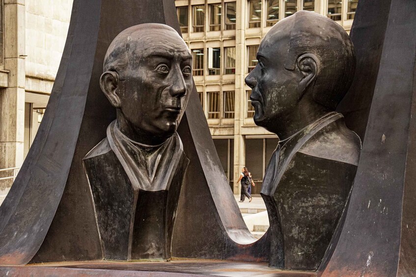 A sculpture of two figures in conversation, set against a building with large windows. 4