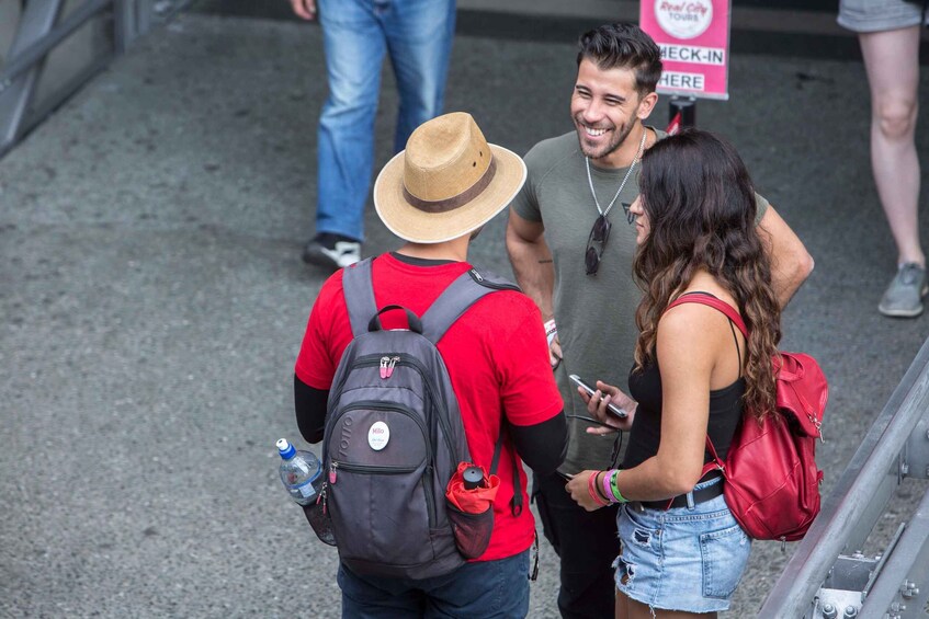 A group of people standing on a sidewalk, some with backpacks and one wearing a hat. 2