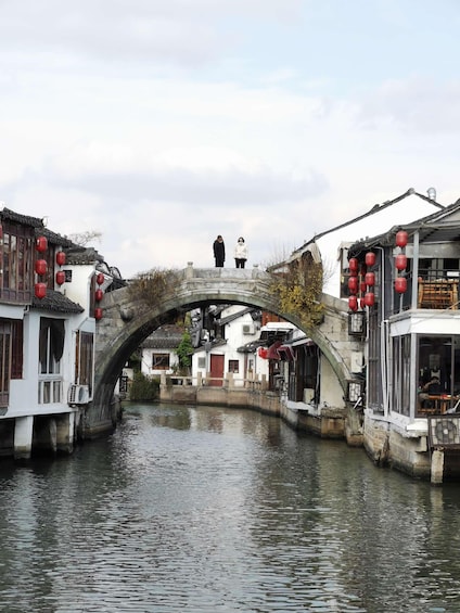 A stone bridge over a canal with traditional buildings on either side, red lanterns, and a person standing on the bridge. 5