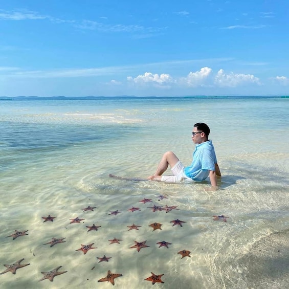 A person sitting in shallow water with starfish on the sand. 1