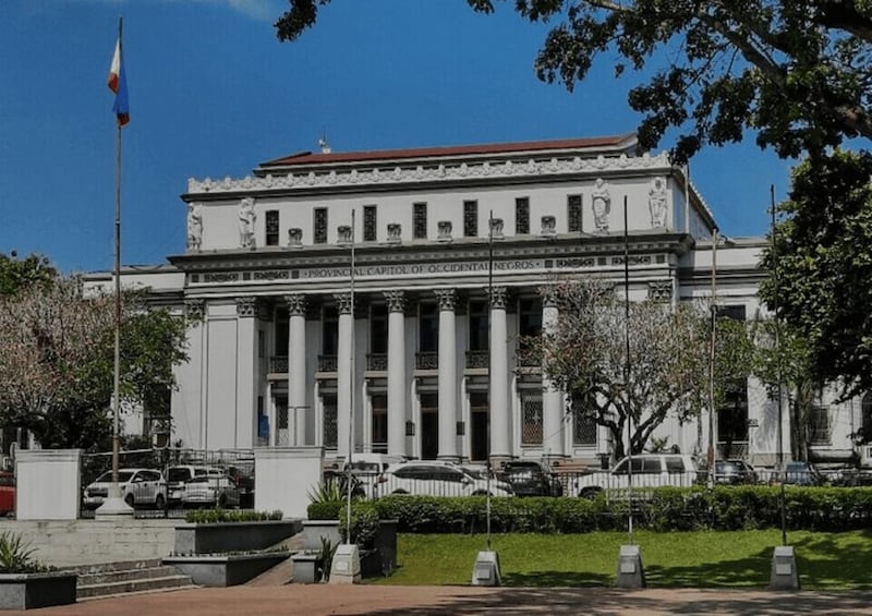 A neoclassical building with columns, a flag, and parked cars. 1