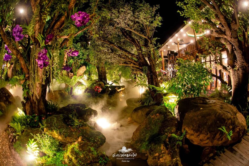 A nighttime garden with a water feature, illuminated plants, and a building in the background. 1