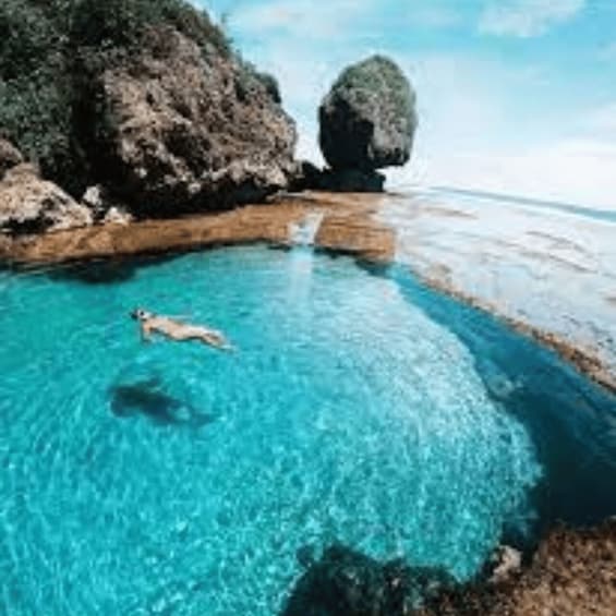 A clear blue pool surrounded by rocky terrain and a large rock formation. 1