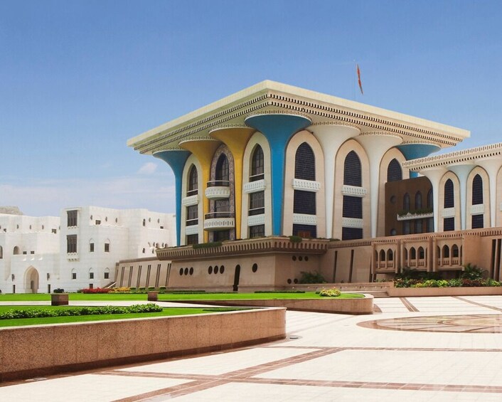 A large, multi-story building with a white facade and blue accents, surrounded by a landscaped courtyard with geometric patterns. 3