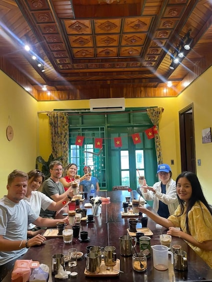 A group of people seated around a table with various drinks and utensils, in a room with wooden paneling and a green door. 2