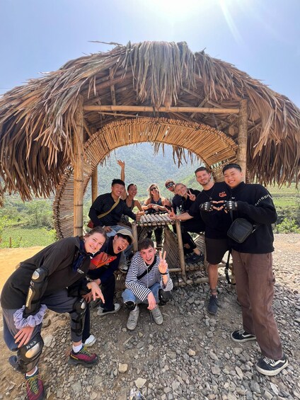 A group of people posing under a thatched structure in a rocky outdoor setting. 2