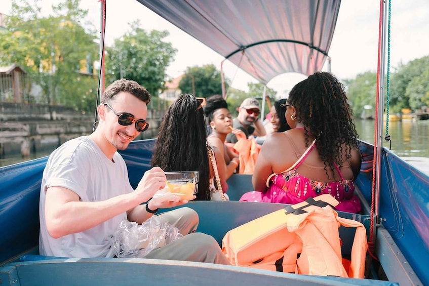 People on a boat with a canopy, some holding drinks, and a river in the background. 1