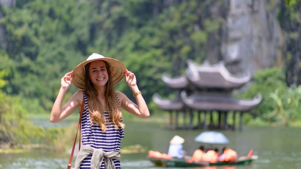 A person in a striped dress and straw hat by a water body with traditional architecture in the background. 1