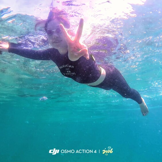 A person underwater making a peace sign, wearing a wetsuit with text, in clear blue water. 5