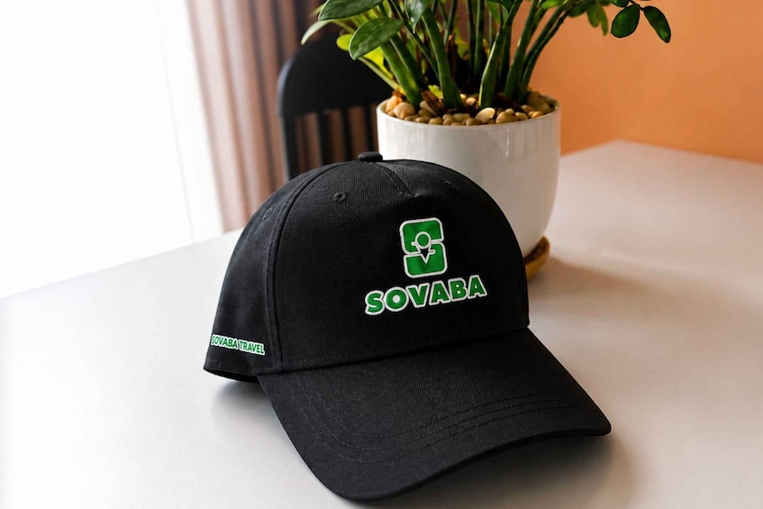 A black cap with 'SOVABA' and 'SOVABATRAVEL' text, a potted plant, and a chair in the background. 4