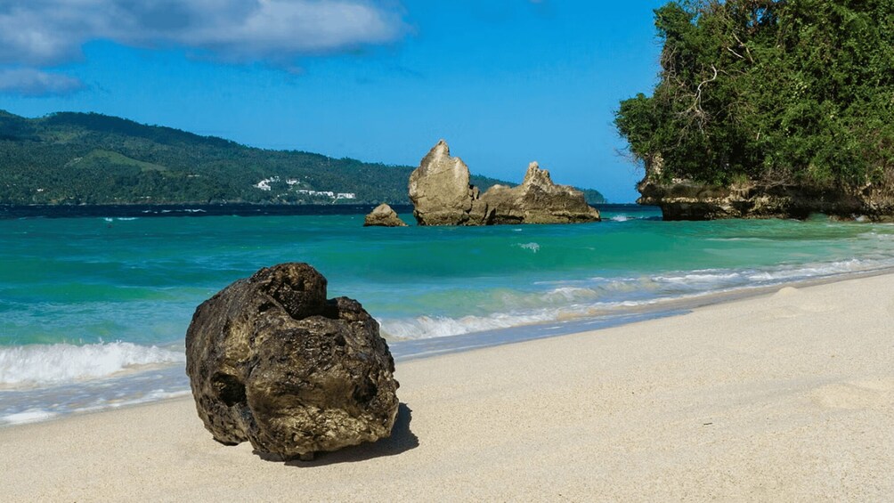 A beach with clear turquoise water, a large rock in the foreground, and rocky outcrops in the distance. 2