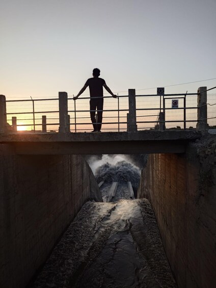 A person standing on a bridge with a railing, overlooking a waterway with a waterfall. 5