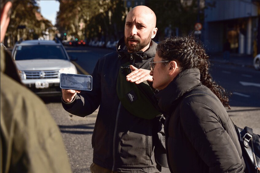 A person holding a smartphone, another person gesturing, and a car in the background. 2