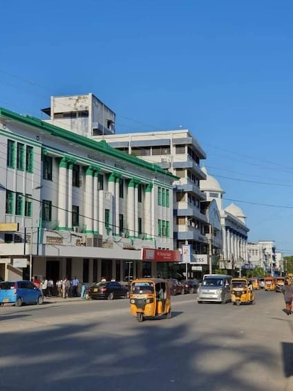 A busy urban street with multi-story buildings, a clear blue sky, and yellow auto-rickshaws on the road. 2