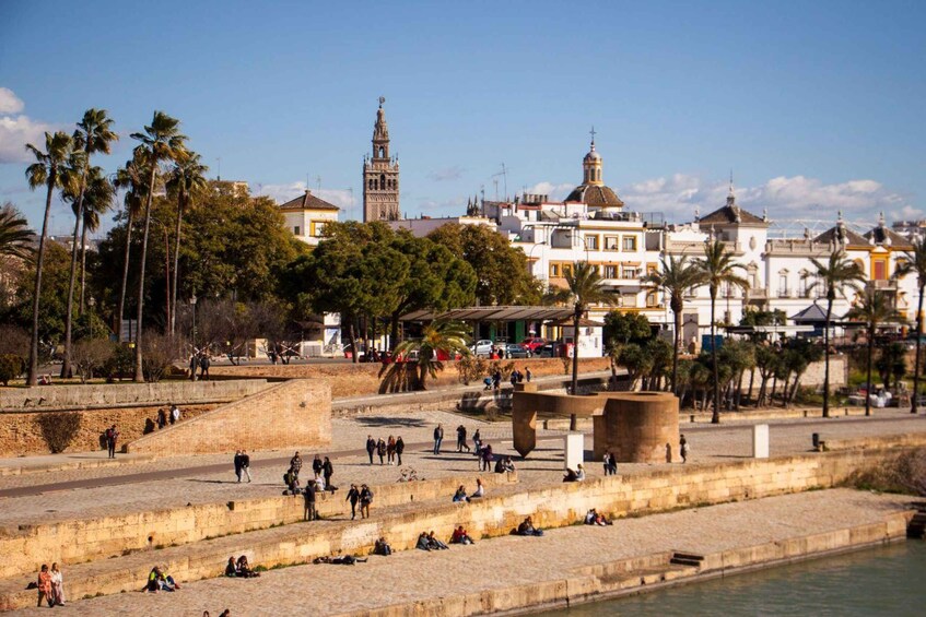 A riverside promenade with people walking and sitting, palm trees, and historic buildings including a prominent tower. 2