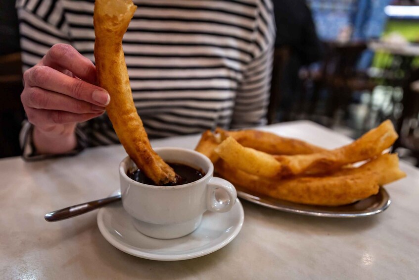A person holding a fried doughstick over a cup of dark liquid, likely coffee, with a plate of similar fried doughsticks beside it. 1