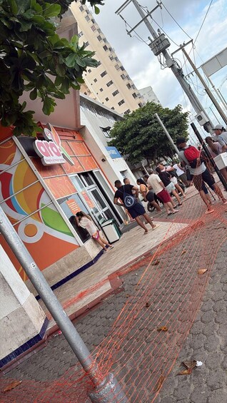 A street scene with a colorful storefront, a tree, and a group of people standing near a fence. 3