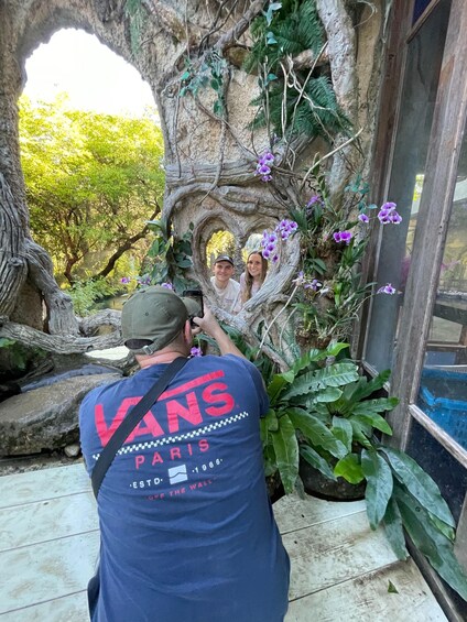 A man taking a photo of two people in a naturalistic setting with a rock arch and purple orchids. 3