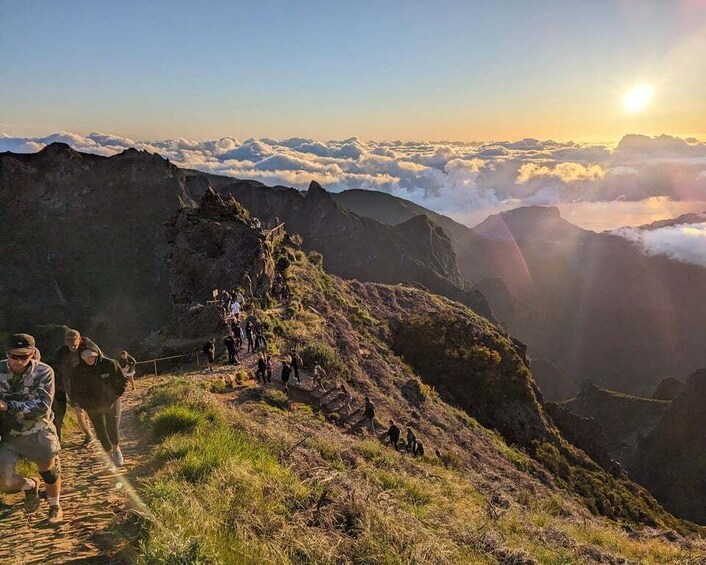 A mountainous landscape with hikers on a trail, surrounded by clouds and the sun setting in the background. 2