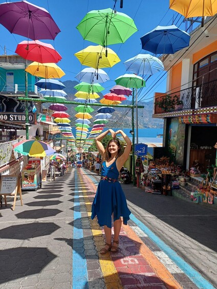 A woman in a blue dress standing on a colorful street with umbrellas overhead. 1