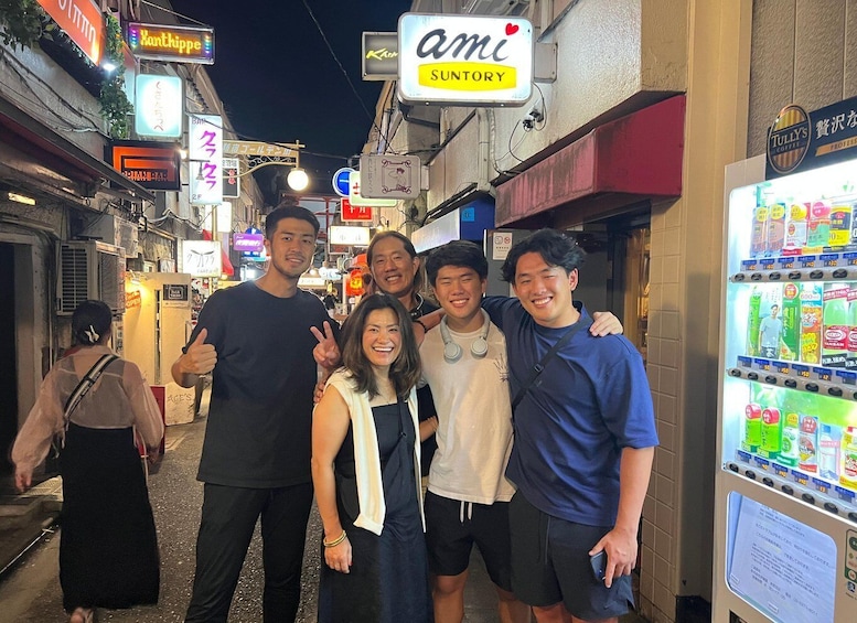 A group of people standing in a narrow street with various illuminated signs and a vending machine. 5