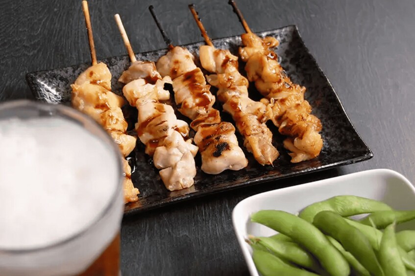 Skewers of grilled chicken on a black plate, a glass of beer, and a bowl of green peas. 2