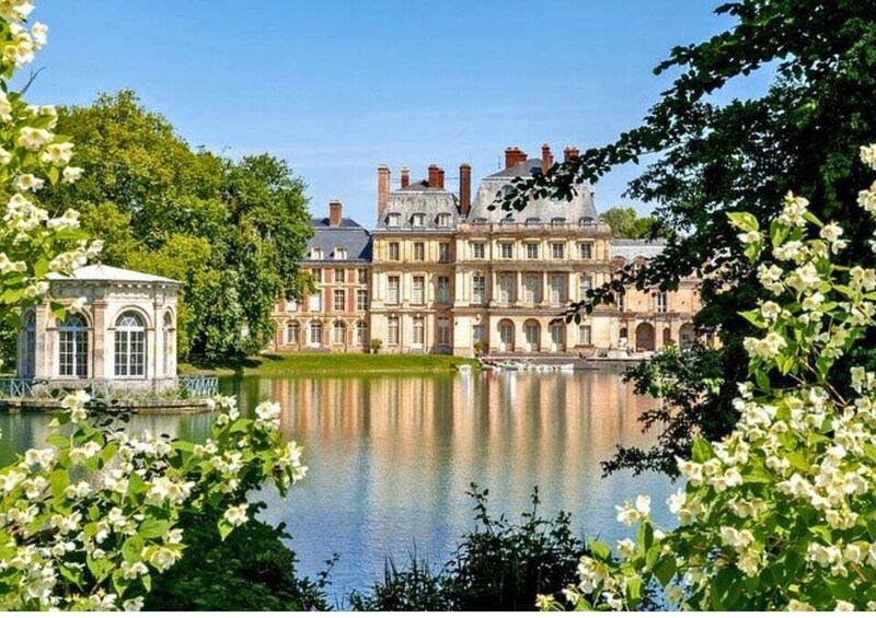 A large, historic building with multiple chimneys and arched windows, surrounded by lush greenery and a calm lake. 1
