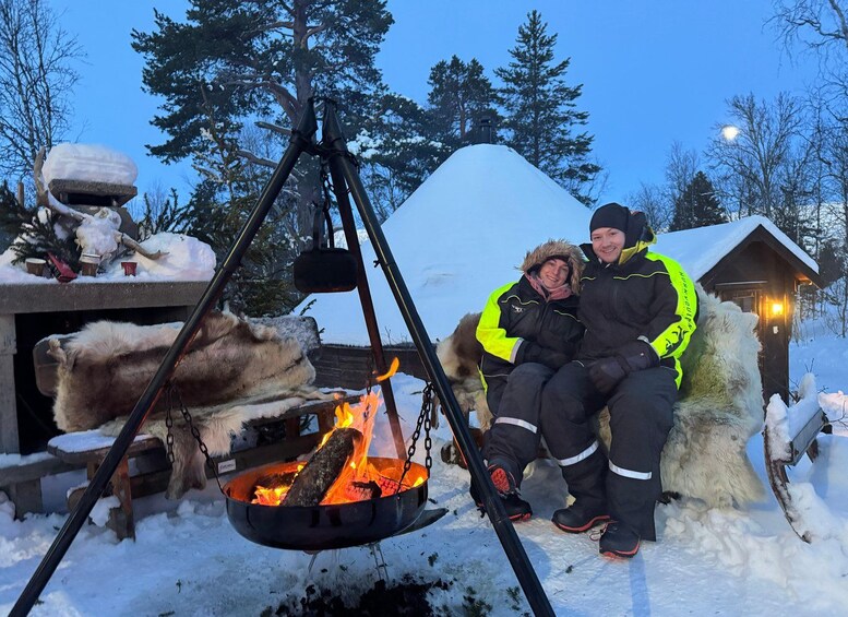 A couple sitting by a campfire with a snow-covered landscape and a wooden cabin in the background. 5