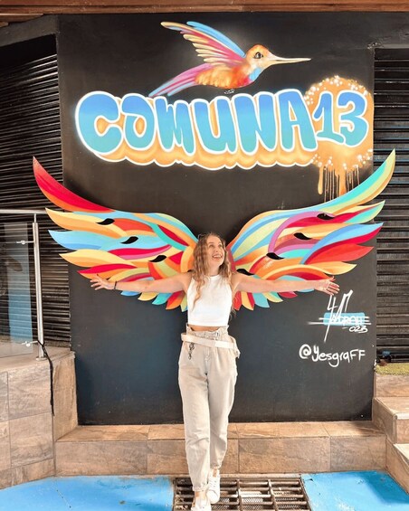 A person standing in front of a mural with a hummingbird and colorful wings, with the text "COMMUNA13" and a hashtag "#4A". 2