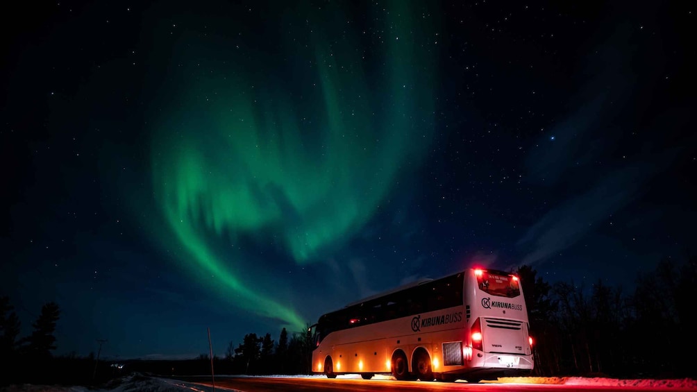 A bus with 'KIRUNABUS' written on it, parked on a road with snow on the sides, under a night sky with the Northern Lights. 1