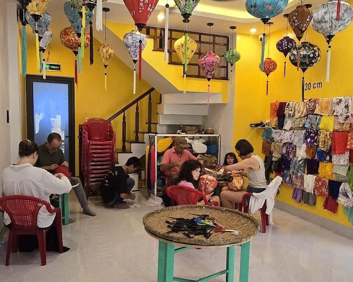 A brightly lit room with yellow walls, hanging lanterns, and a staircase. People are seated and working at a table with various items. 5