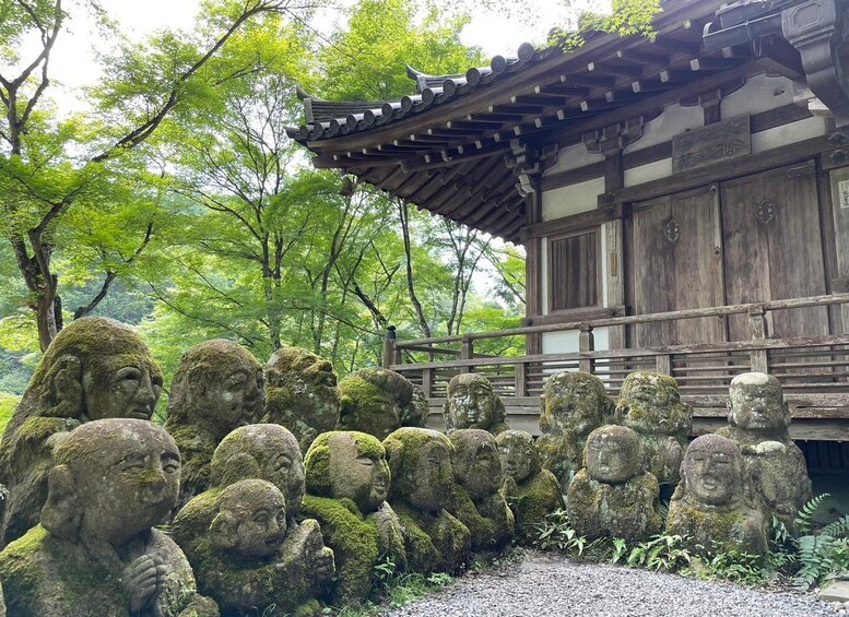 A traditional Japanese building with a tiled roof and wooden walls, surrounded by a forest. In front, there is a stone arrangement of Buddha statues. 1
