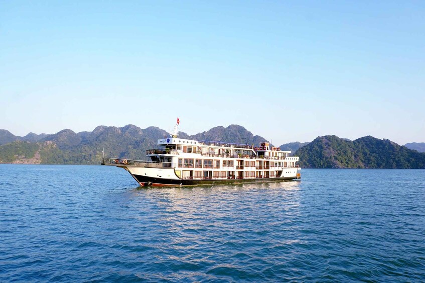 A white riverboat with multiple decks and a red flag on top, floating on calm water with mountains in the background. 1