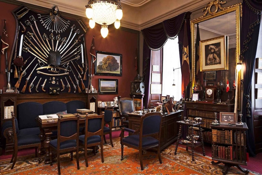 A room with a large collection of antique swords displayed on a wall, a chandelier, a large mirror, and a red carpet. 4