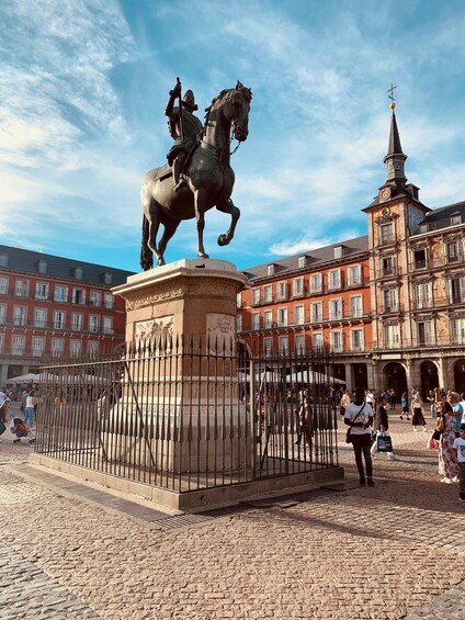 A statue of a man on a horse in a square with cobblestone pavement, surrounded by historic buildings and a clock tower. 5