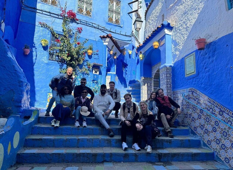 A group of people sitting on blue-painted stairs in a narrow alleyway with decorative tiles and hanging plants. 5