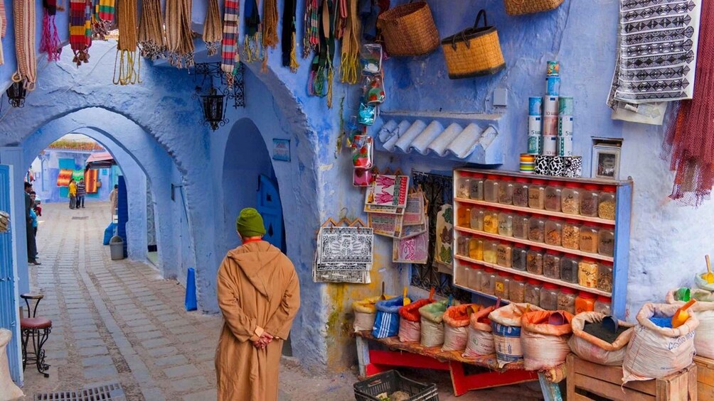 A narrow alleyway with a man in traditional attire, colorful textiles, and various goods displayed on shelves and hanging from the walls. 1
