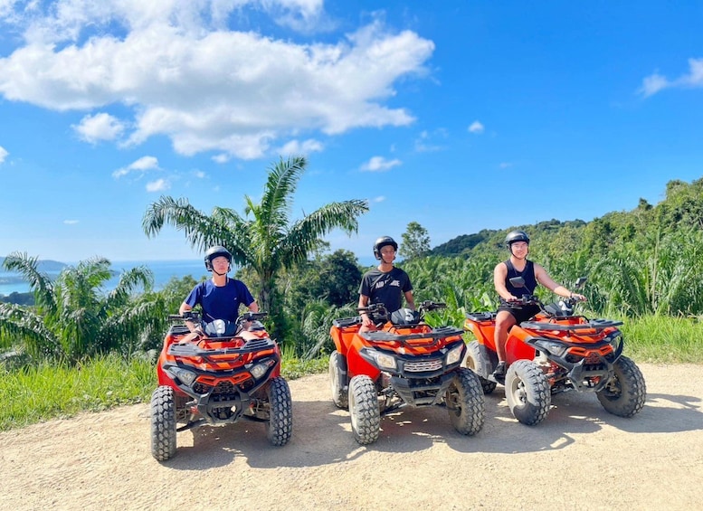 Three people on ATVs on a dirt path with a tropical landscape and palm trees in the background. 2