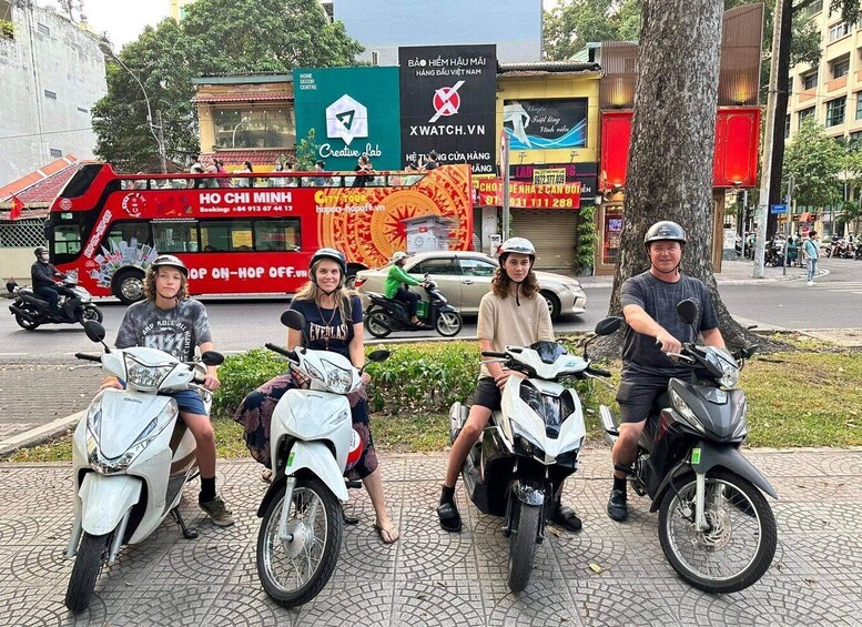 Four people on scooters in an urban setting with commercial buildings and a red bus in the background. 2