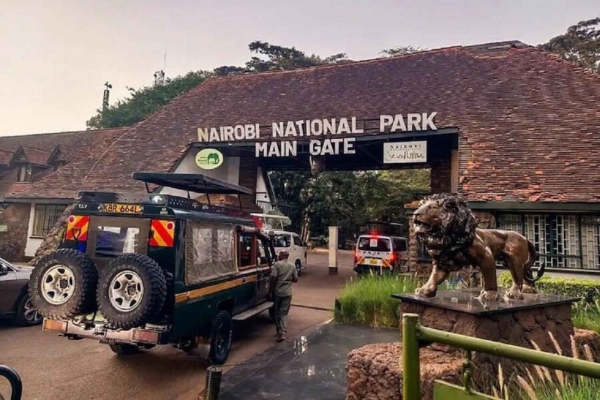 The Nairobi National Park main gate with a lion statue and vehicles parked in front. 1