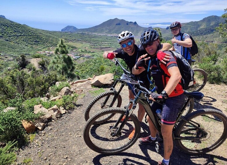 Three people on mountain bikes on a dirt trail with a scenic view of mountains and greenery in the background. 1