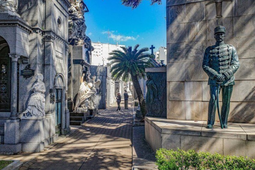 Une statue d’un soldat en uniforme, un bâtiment en pierre à l’architecture classique et un palmier dans un environnement urbain ensoleillé. 1
