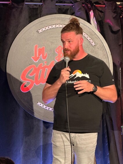 A person holding a microphone, wearing a black t-shirt with a mountain design and the word 'Columbia' on it, standing in front of a backdrop with a circular design and the words 'In the' visible. 2