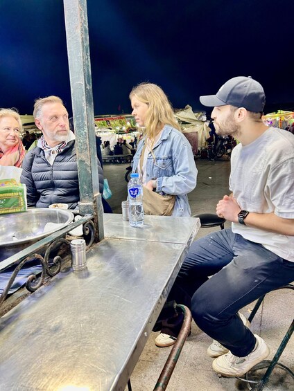 A group of people at an outdoor market with food stalls and a metal table in the foreground. 3