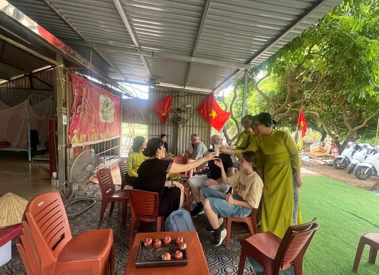A group of people seated at a table under a covered area with flags, surrounded by trees and parked scooters. 4