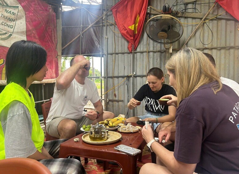 A group of people sitting at a table with food and a fan in a makeshift shelter. 5
