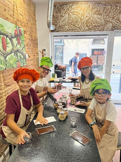 Four people wearing chef hats and aprons, sitting at a table with jars of ingredients and chocolate bars. 2