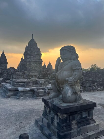 A stone statue of a seated figure with a large belly, surrounded by stone blocks and a temple in the background. 1