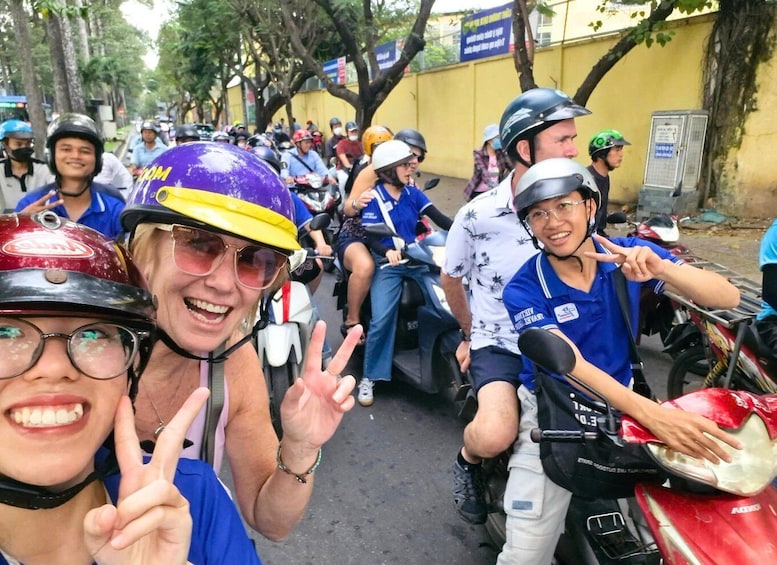 A group of people on motorcycles, some wearing helmets and casual clothing, gathered on a street with trees and buildings in the background. 1